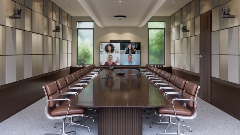 Modern boardroom with a long table, leather chairs, wall mounted cameras, ceiling microphones, and a screen showing a four-person video conference call.