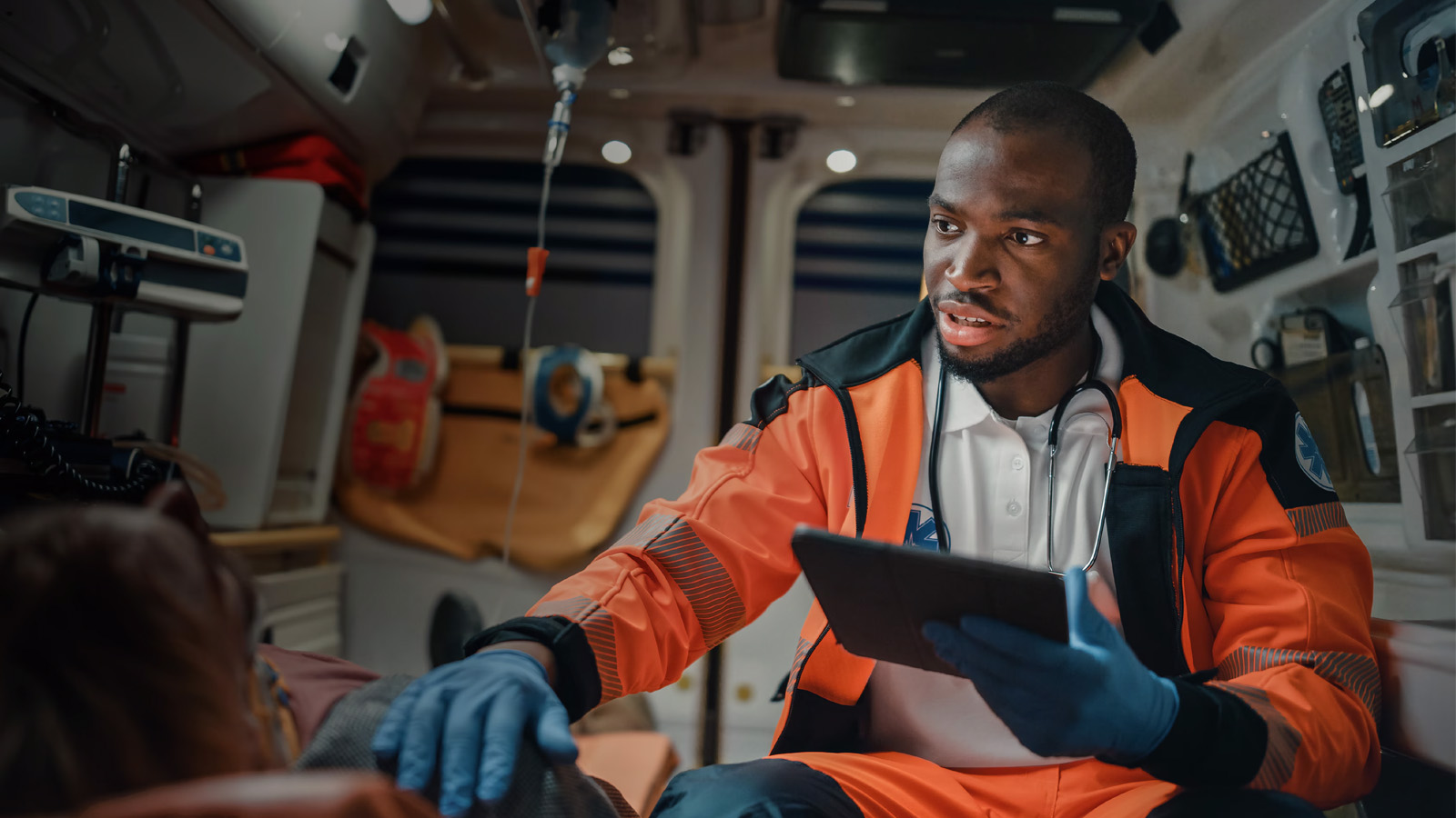 A first responder in an ambulance uses a tablet while resting a hand on a patient's shoulder.