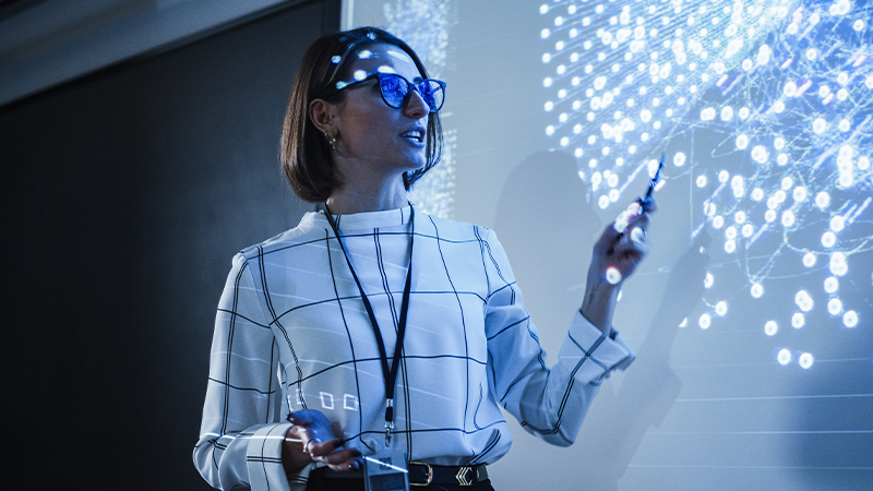 Professional woman presenting complex data visualization and network analytics on a large digital screen.