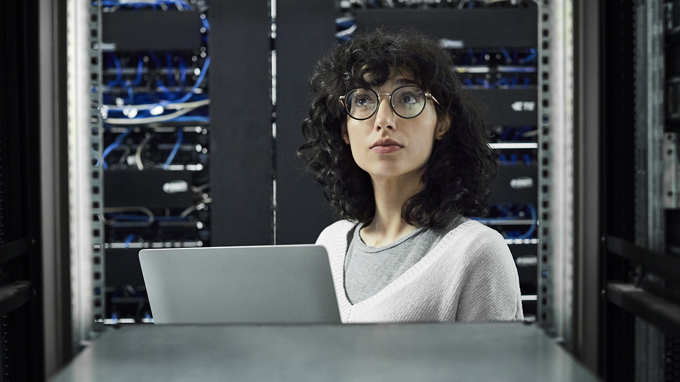 Female technician looking at cables. Engineer with laptop standing at workplace. She is in server room.